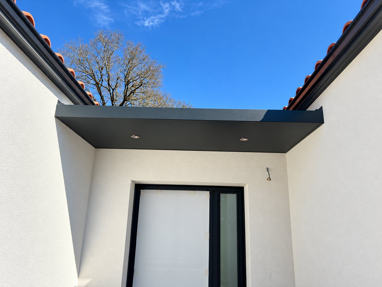 Auvent plat gris anthracite équipé de spots encastrés au-dessus d'une porte-fenêtre à cadre noir, sur une façade blanche enduite et sous un ciel bleu.