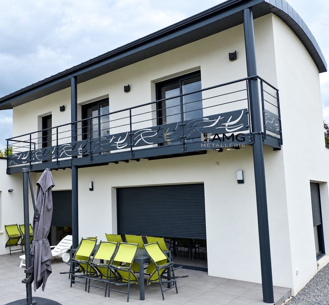 Maison moderne à étage dont le balcon est équipé d'un garde-corps en métal anthracite ajouré, au-dessus d'une terrasse avec chaises vert anis et parasol fermé.