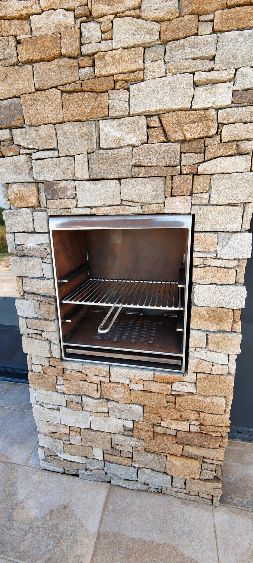 Un barbecue en inox avec grille est encastré dans un muret en pierres naturelles, au-dessus d'un sol dallé en extérieur.