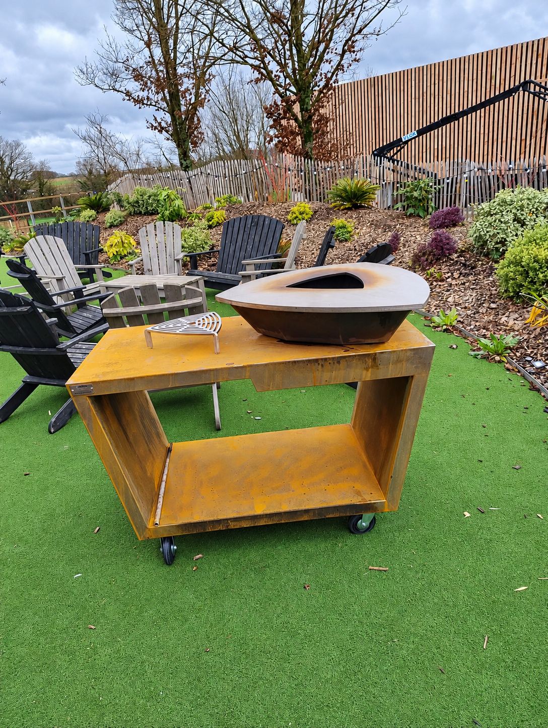 Un brasero triangulaire en acier corten posé sur un chariot à roulettes, sur une pelouse synthétique devant des fauteuils Adirondack et un massif planté.