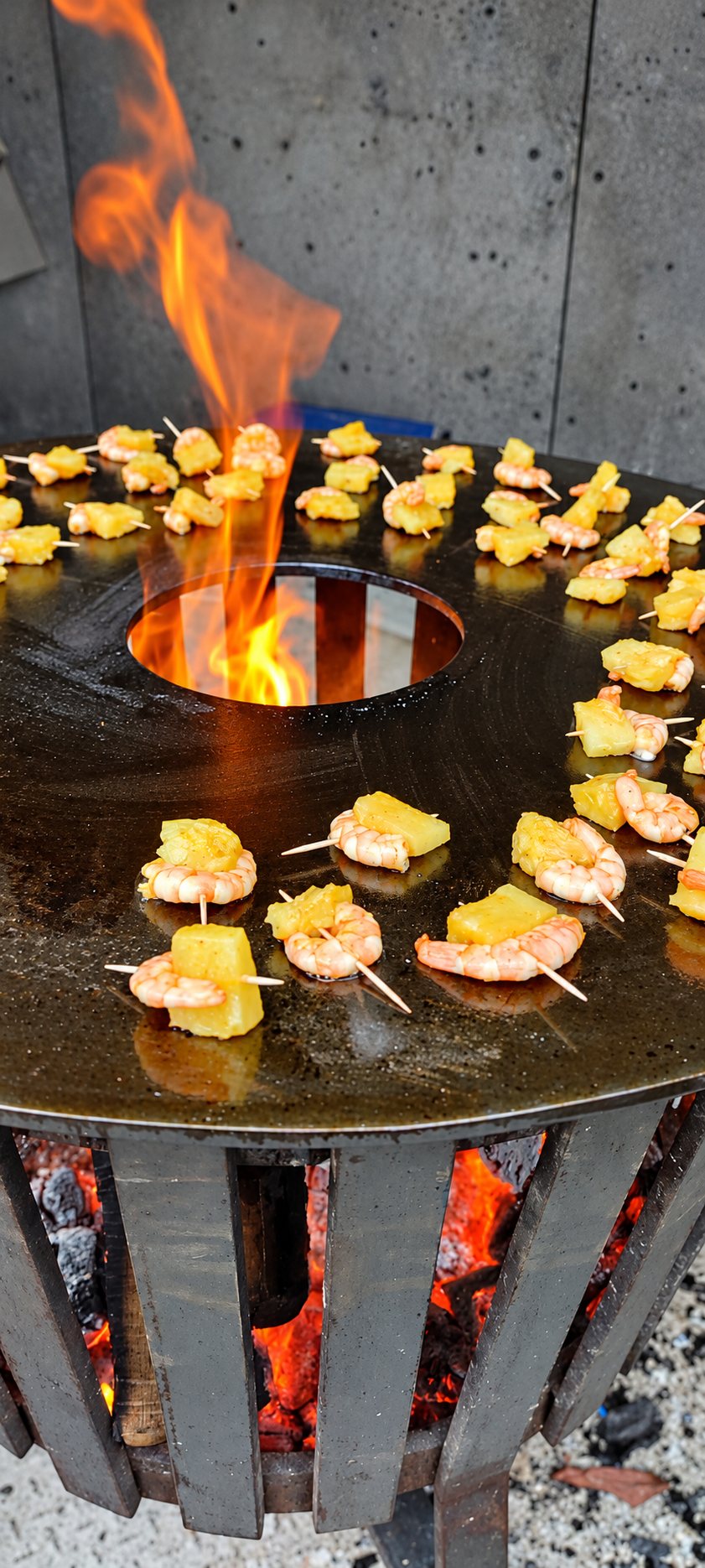 Des brochettes de crevettes et de morceaux d'ananas cuisent sur une plancha ronde en acier chauffée par un feu de bois dont les flammes sortent par le centre.