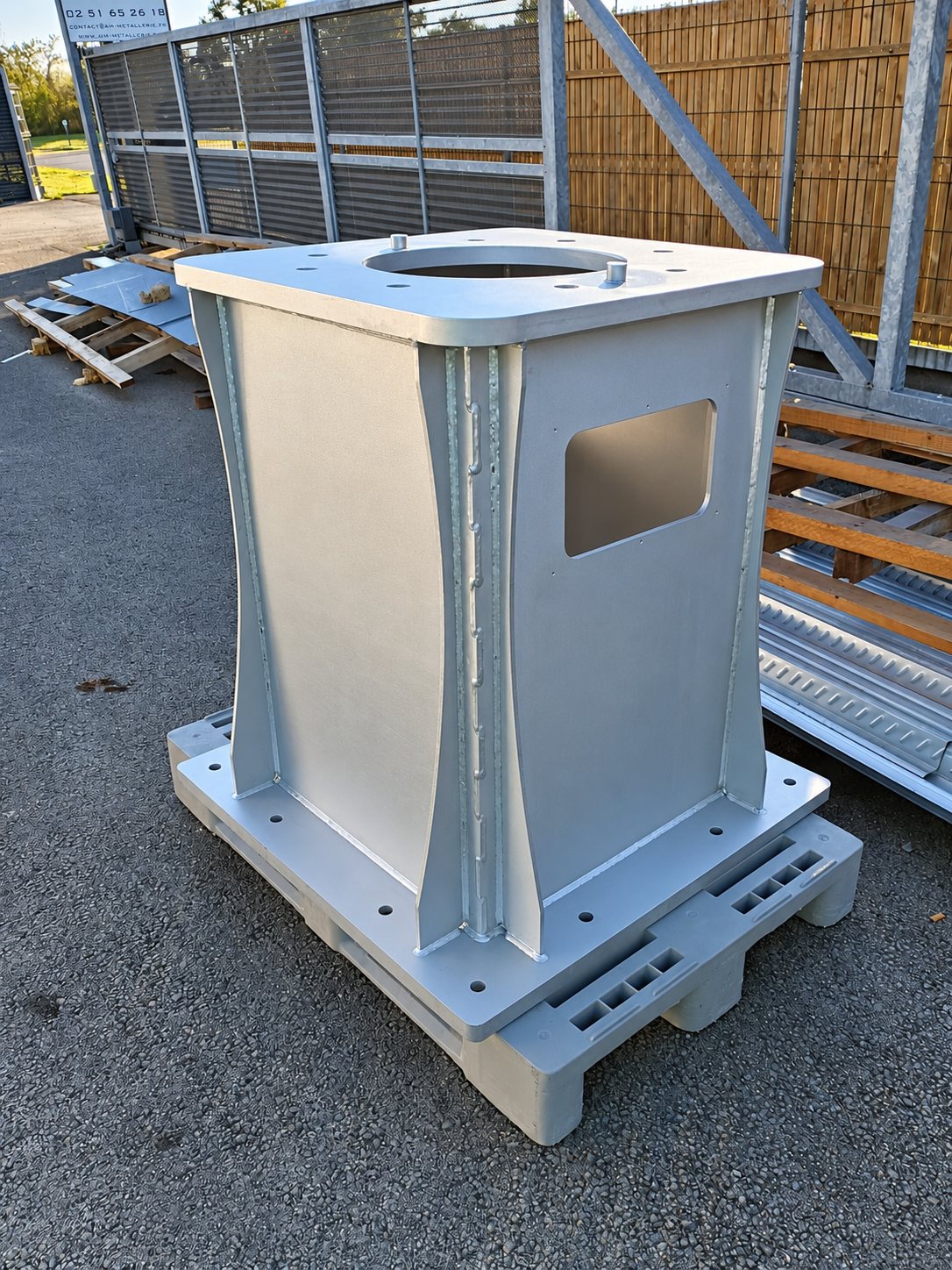 Un caisson en acier soudé, avec platines percées et ouverture latérale, posé sur une palette en plastique sur un parking, devant une clôture.