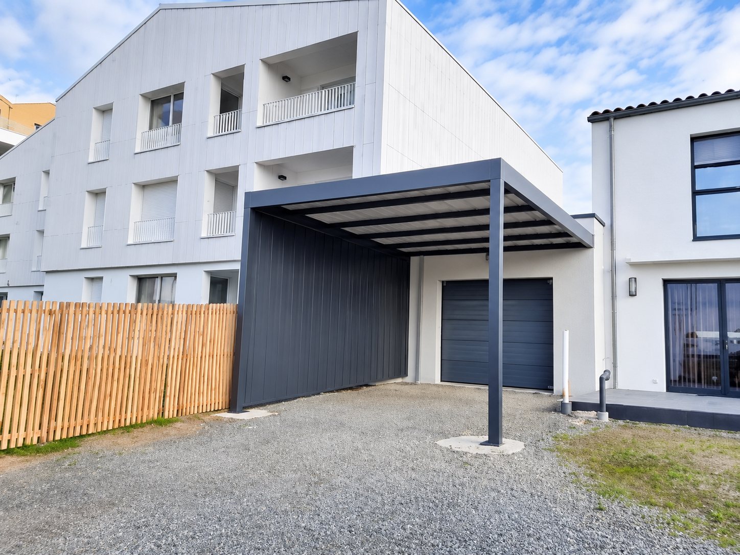 Un carport moderne à toit plat et panneaux gris foncé est installé sur une allée de gravier devant un bâtiment contemporain blanc et gris, à côté d'une clôture en bois.