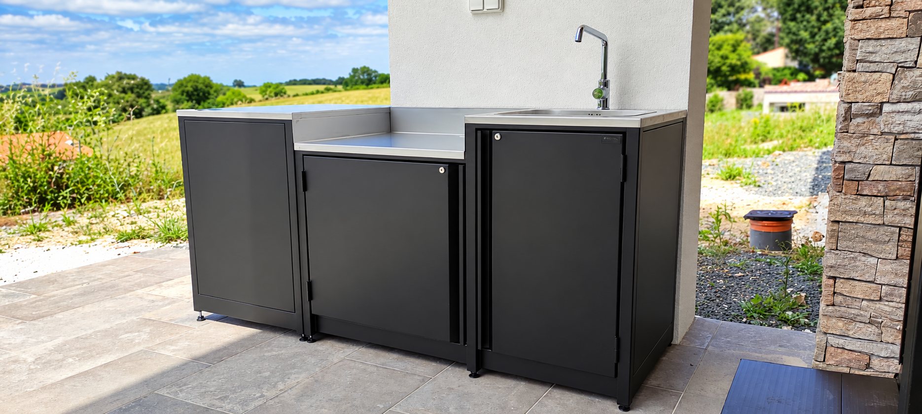 Un module de cuisine d'extérieur en métal noir avec plan de travail et évier en inox est installé sur une terrasse dallée, face à un paysage de campagne.