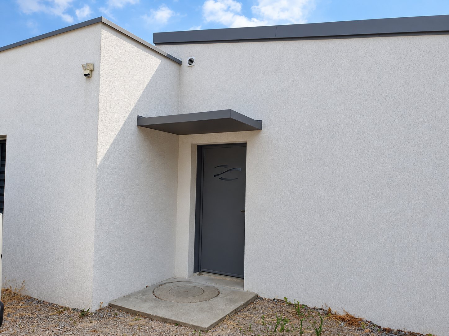 Porte d'entrée gris anthracite protégée par un auvent métallique, sur la façade enduite blanche d'une maison moderne à toit plat.