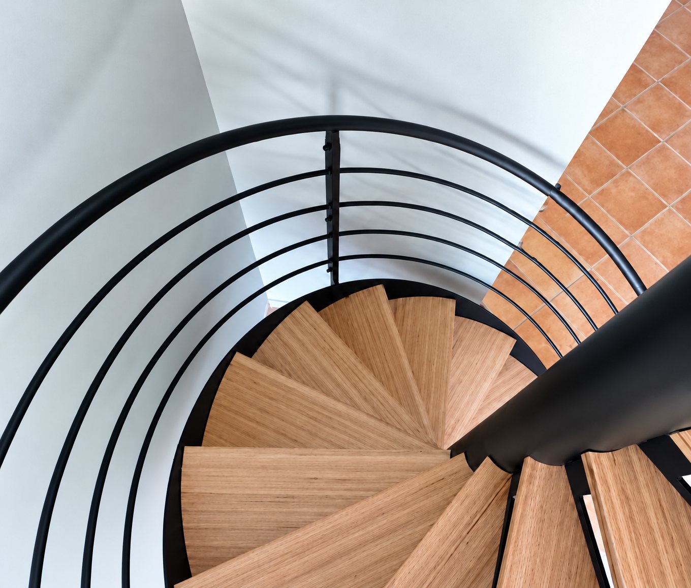 Vue du dessus d’un escalier en colimaçon aux marches en bois clair, entouré d’une rampe métallique noire à lisses.