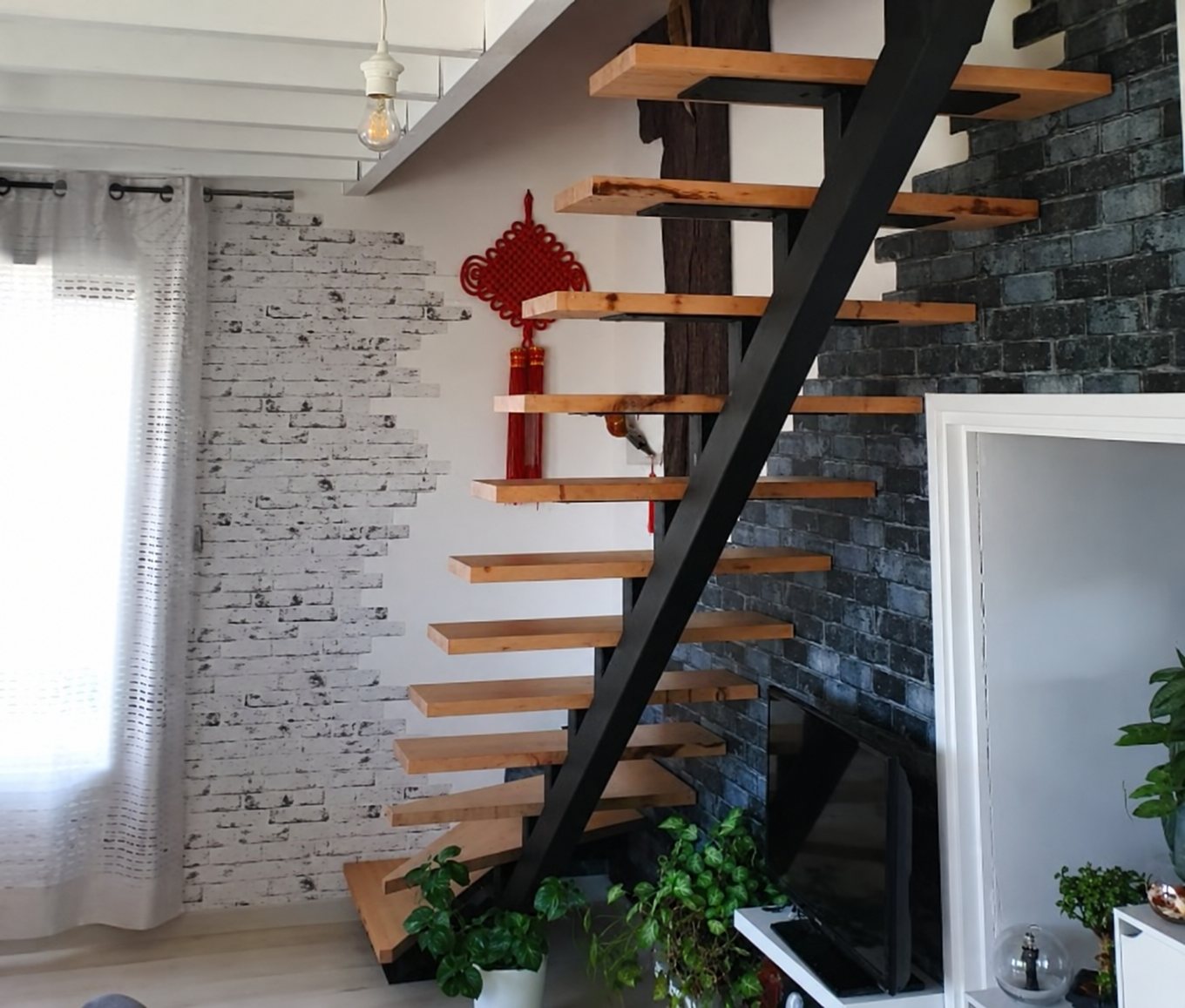 Escalier suspendu à limon central en acier noir et marches en bois, près d’un mur de briques.