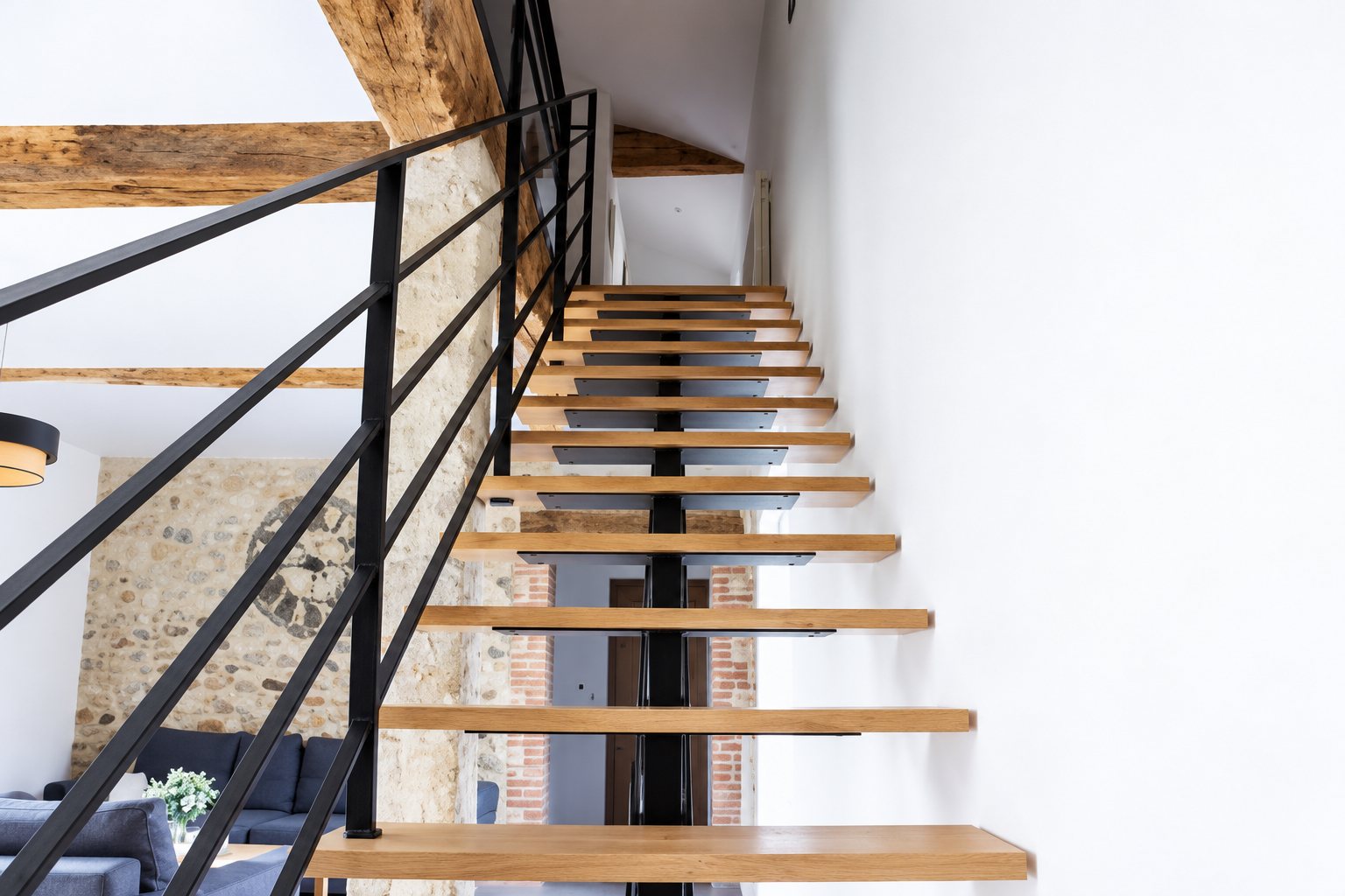 Escalier intérieur aux marches en bois clair et à la rampe en métal noir, entouré de murs blancs, de pierres apparentes et d'une poutre ancienne.