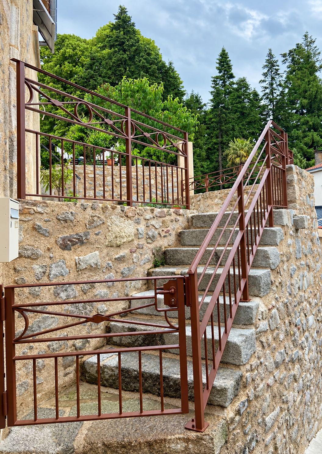 Un escalier en pierre de granit bordé de rampes et d'un portillon en métal brun, avec des arbres en arrière-plan.
