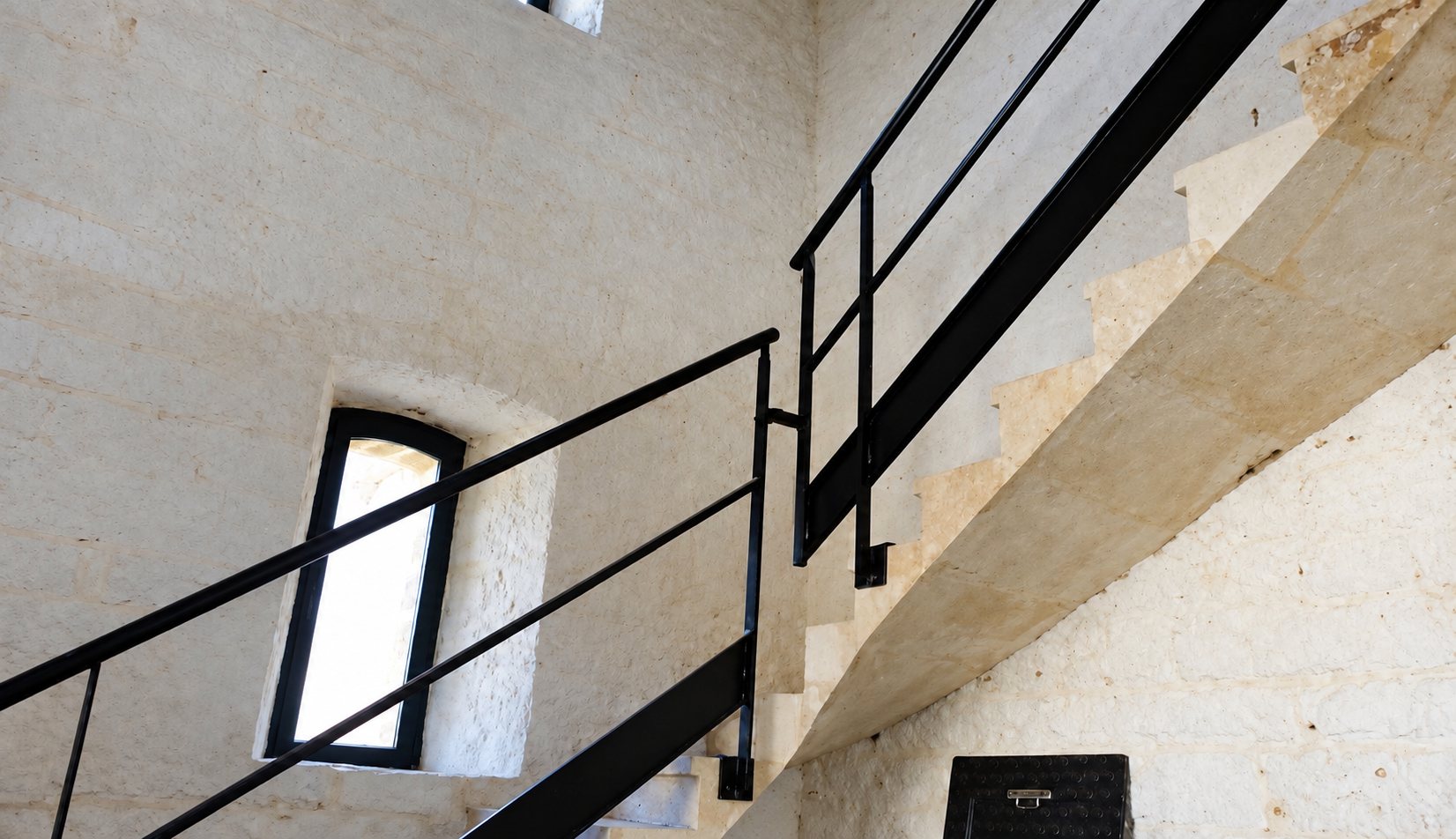 Un escalier aux marches claires et à la rampe métallique noire monte le long d’un mur de pierre brute, près d’une petite fenêtre cintrée.