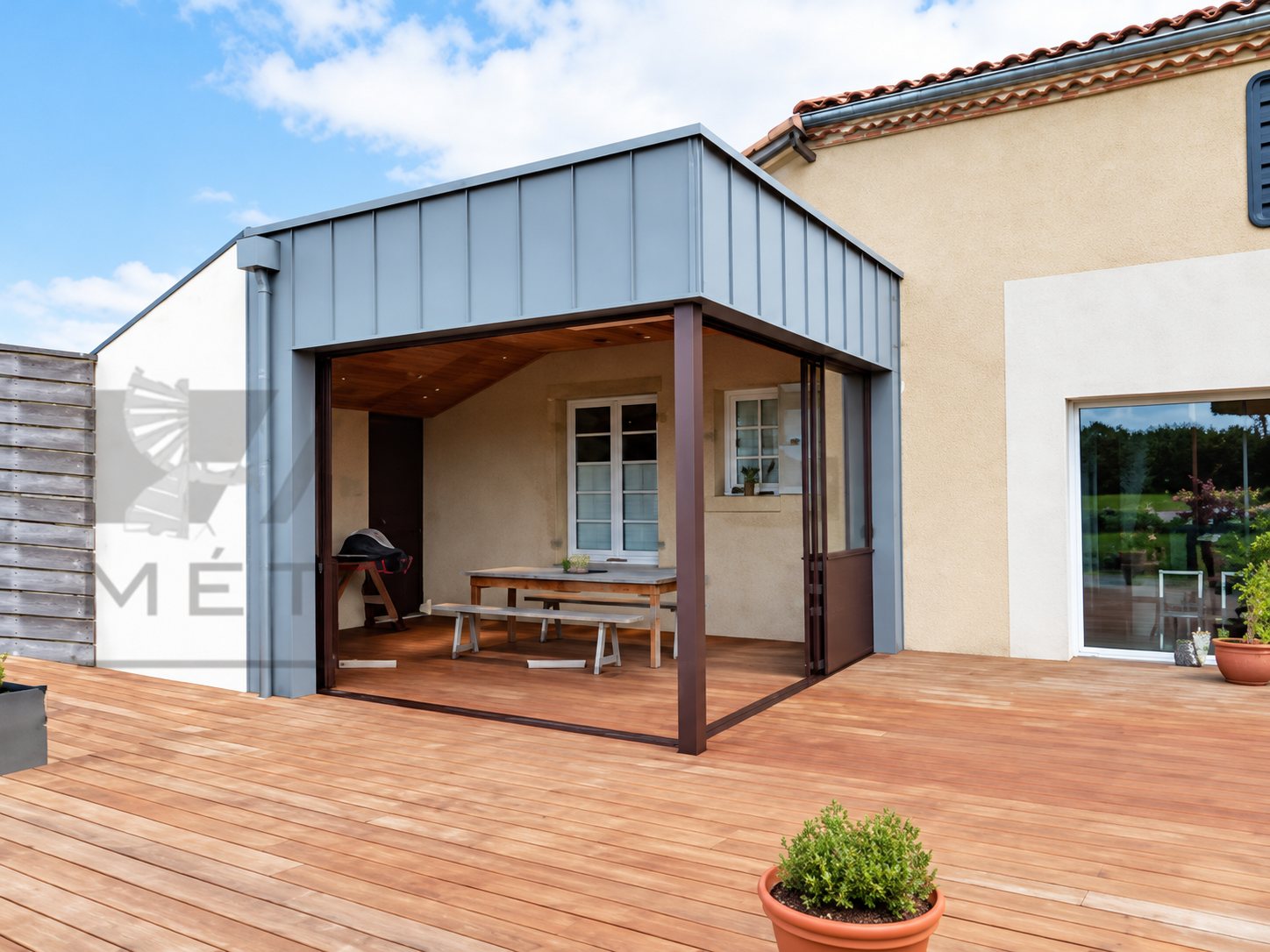 Une extension à toit plat avec bardage métallique gris-bleu ouvre sur une terrasse en bois devant une maison en crépi beige.