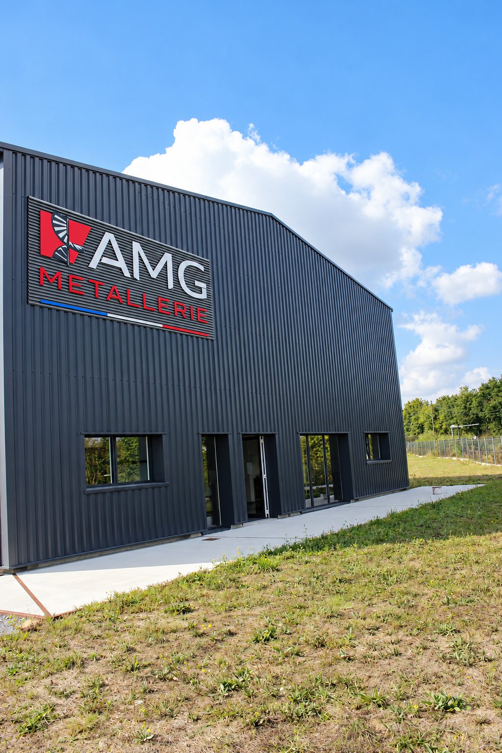 Bâtiment industriel en bardage métallique gris foncé portant l'enseigne AMG Métallerie, avec un trottoir en béton et une pelouse au premier plan.