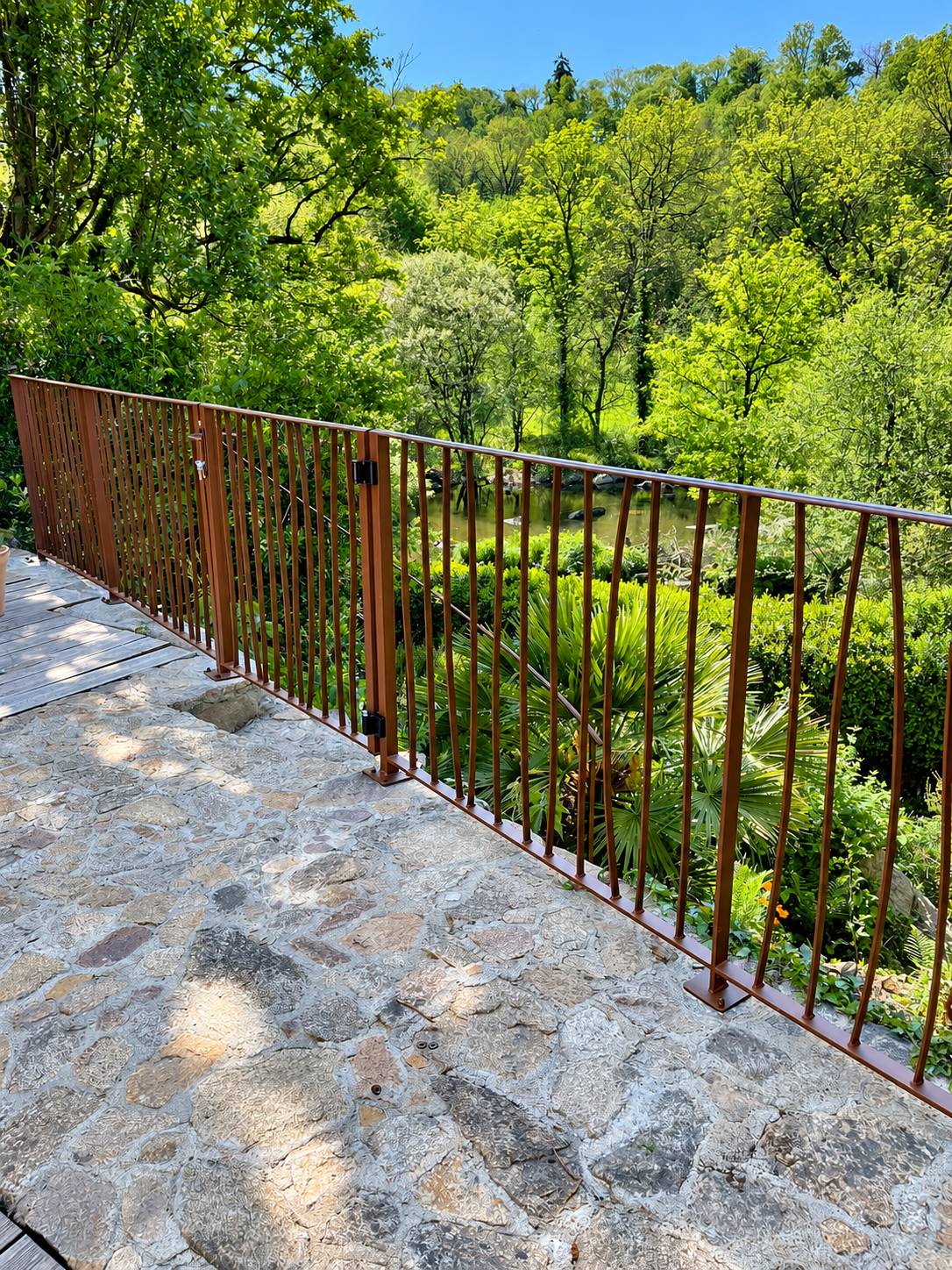 Un garde-corps en métal brun à barreaux verticaux borde une terrasse en pierre qui domine un étang entouré d'arbres verts.