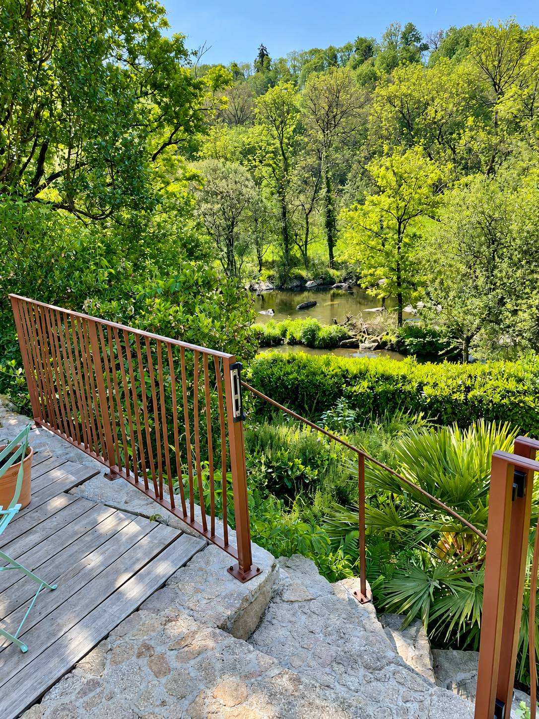Un garde-corps en métal brun longe des marches en pierre et une terrasse en bois, avec vue sur une rivière entourée d'arbres verts.