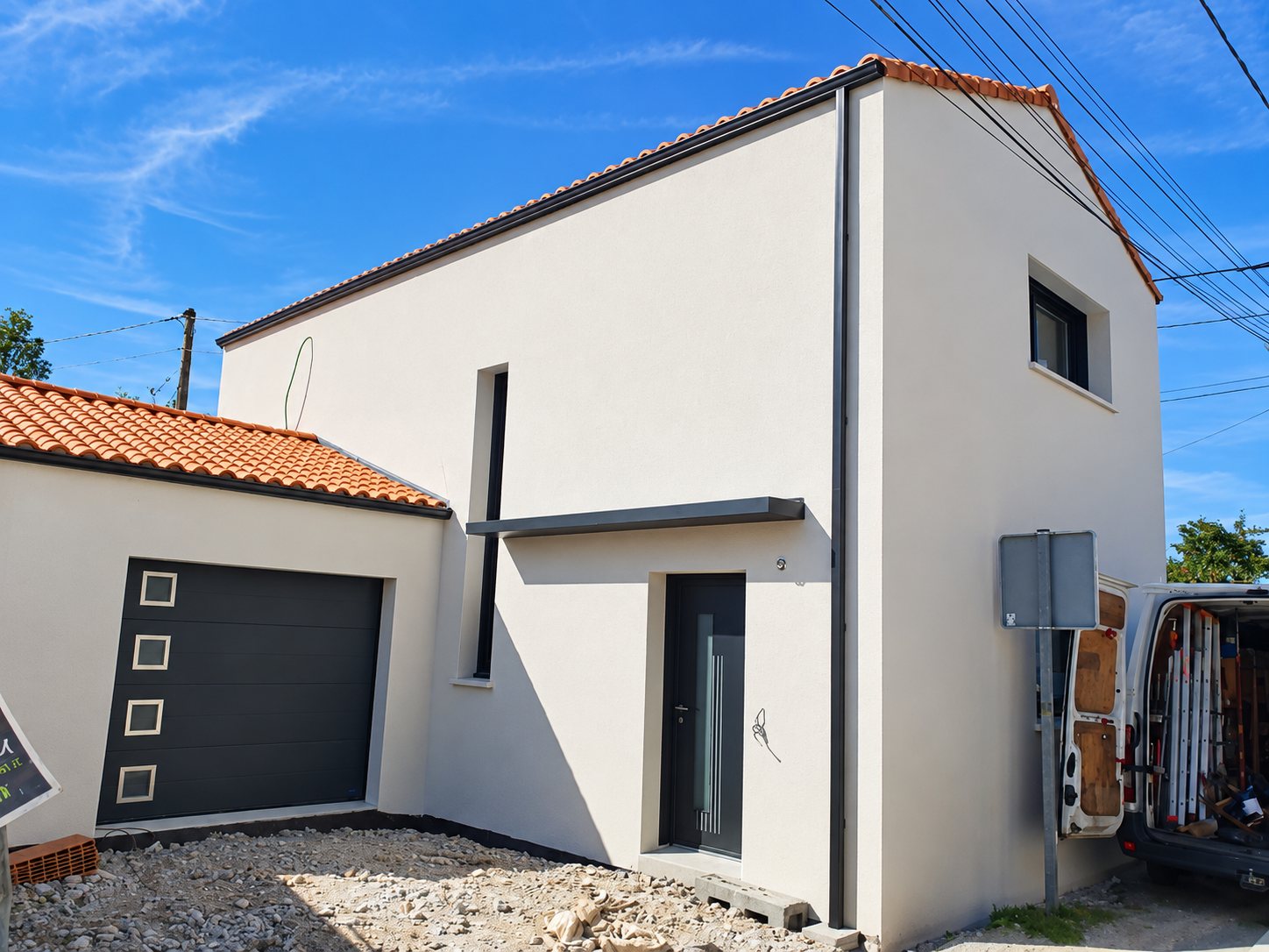 Maison récemment construite à façade blanc cassé, avec porte de garage et porte d'entrée gris anthracite, et une camionnette d'artisan ouverte garée à droite.