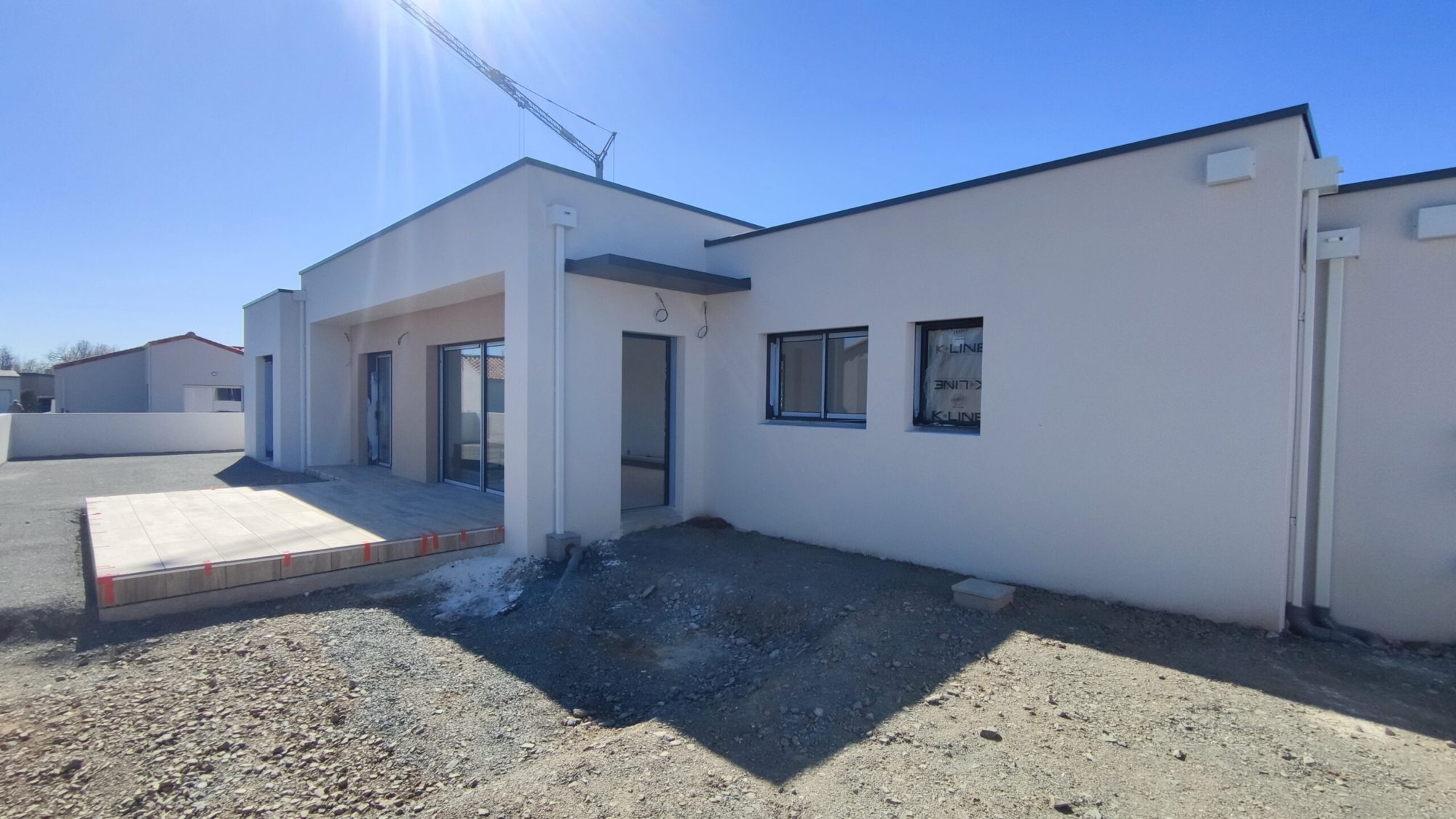 Maison contemporaine de plain-pied aux murs blancs et à toit plat, avec une terrasse carrelée en cours de réalisation sur un terrain de chantier, sous un ciel bleu.