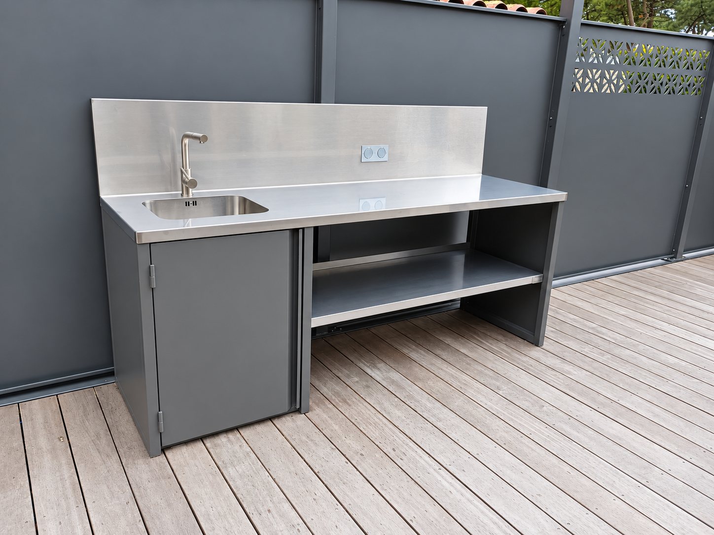 Un meuble d'extérieur en inox et métal gris anthracite avec évier, robinet et étagère basse, installé sur une terrasse en bois devant des panneaux gris.