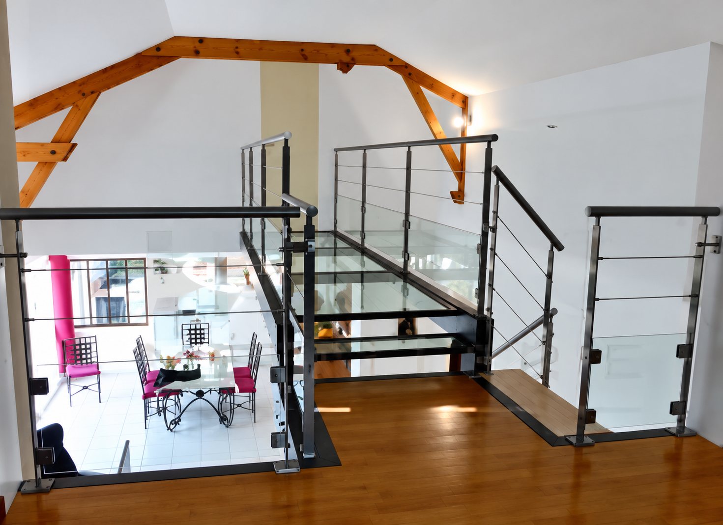 Mezzanine lumineuse avec parquet, garde-corps en inox à câbles et passerelle vitrée, sous une charpente en bois apparente.