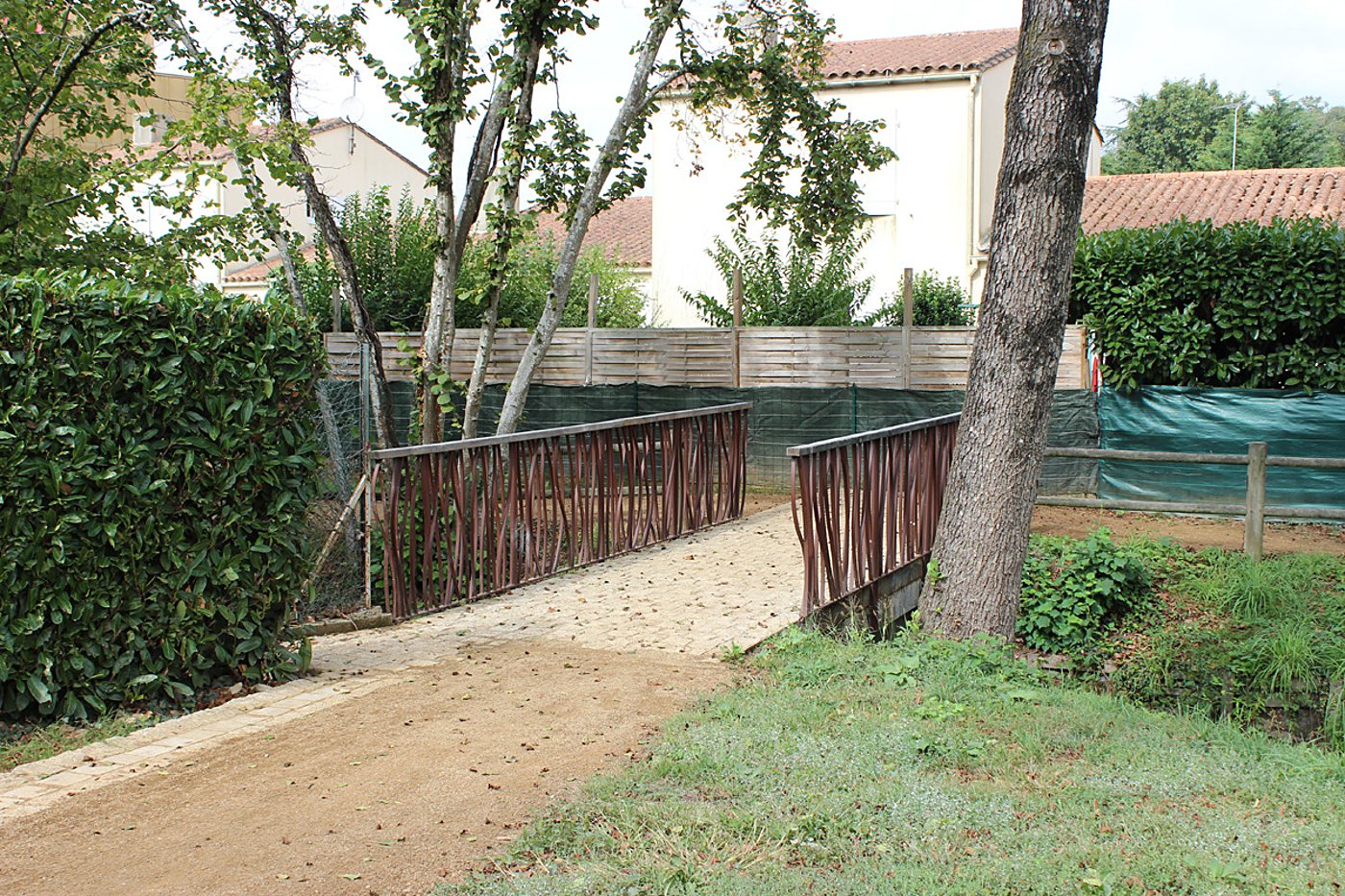 Une petite passerelle pavée avec des garde-corps en bois sculpté relie deux chemins de gravier dans un jardin entouré de haies et de maisons.