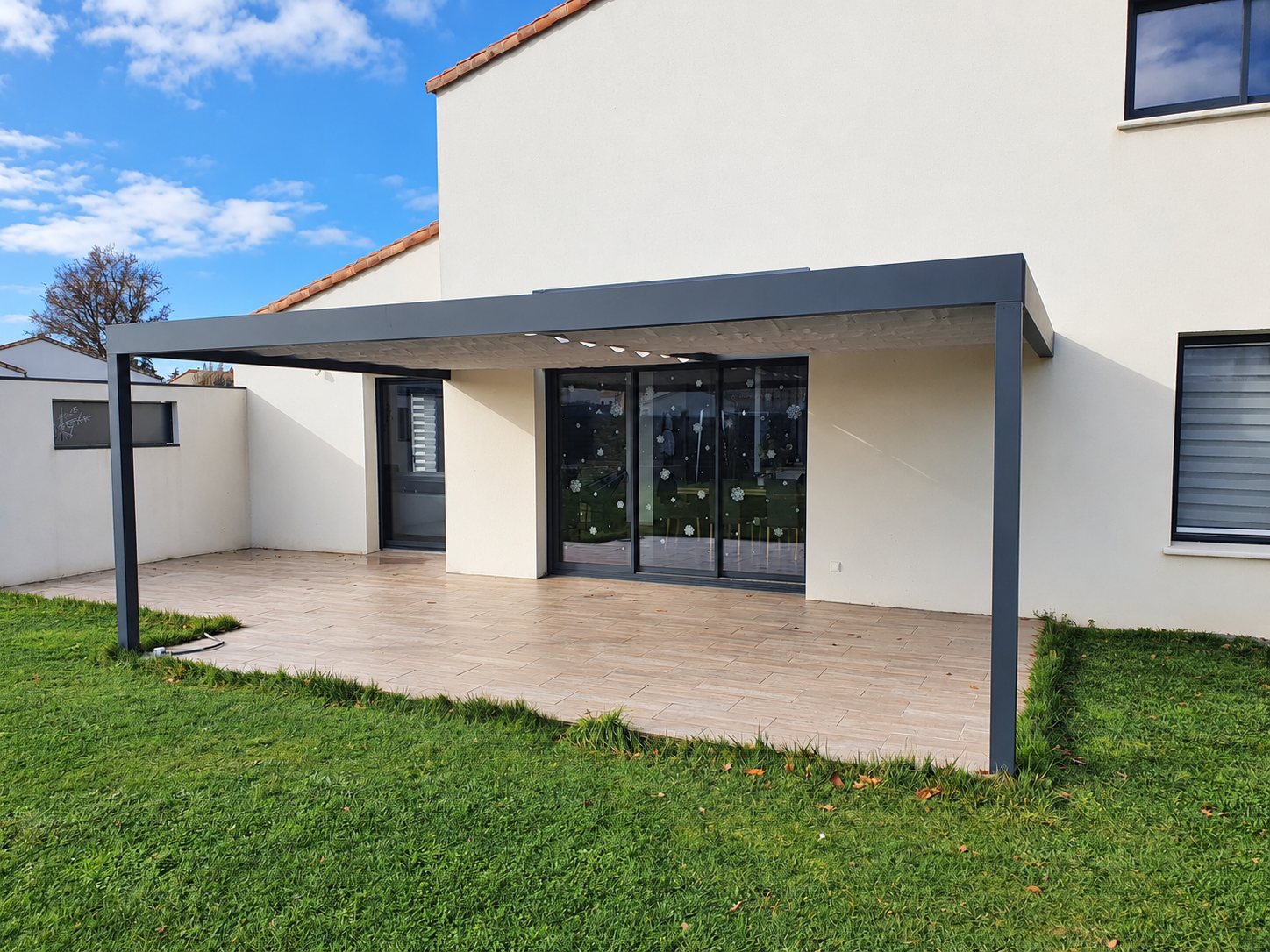 Une pergola en aluminium gris anthracite couvre une terrasse carrelée effet bois devant les baies vitrées d'une maison blanche, avec une pelouse au premier plan.