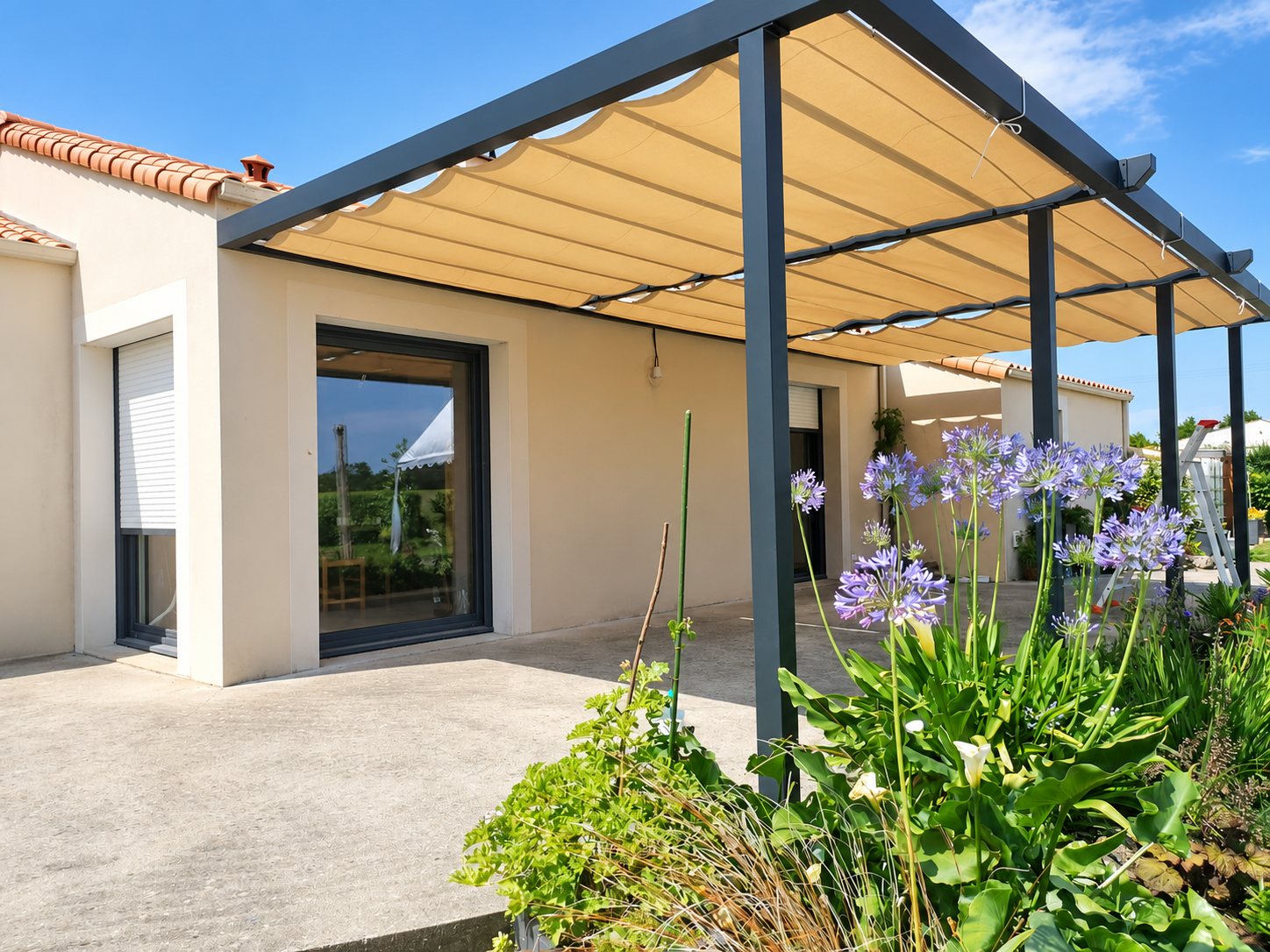 Pergola à structure grise et toile coulissante beige installée sur la terrasse d'une maison, avec des agapanthes en fleurs au premier plan.