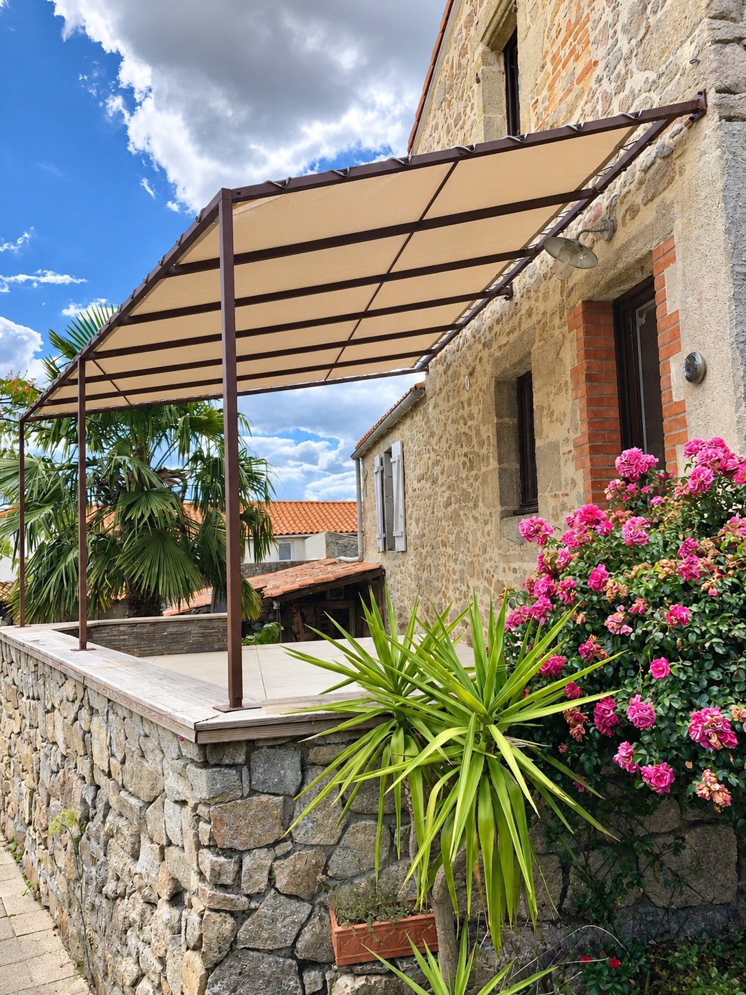 Une pergola en métal marron avec toile beige couvre une terrasse en pierre à côté d'une maison ancienne et d'un rosier fleuri.