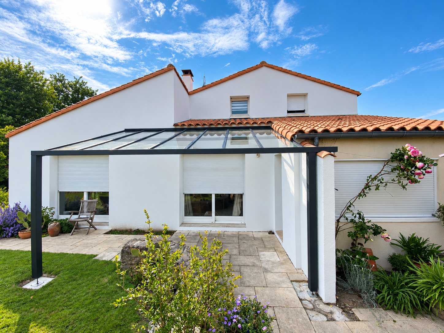 Une pergola en aluminium anthracite avec toit en verre est installée contre une maison blanche à toit de tuiles, devant une terrasse dallée et un jardin fleuri.