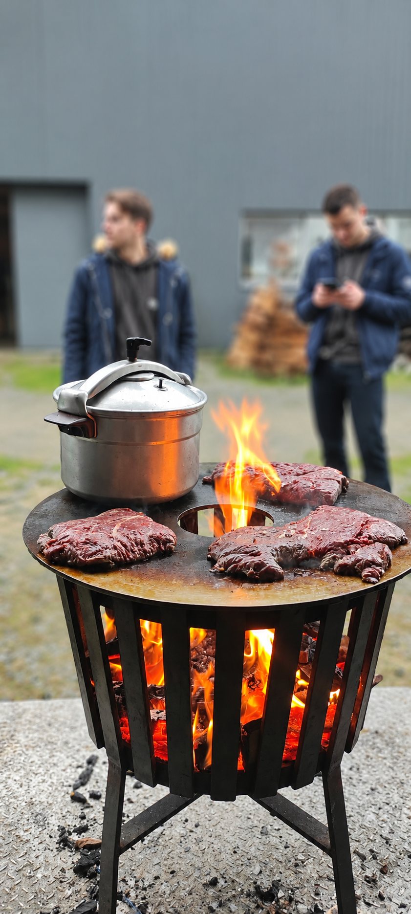 Un brasero en acier surmonté d'une plancha ronde où cuisent trois steaks et où est posé un autocuiseur, avec deux personnes floues à l'arrière-plan.
