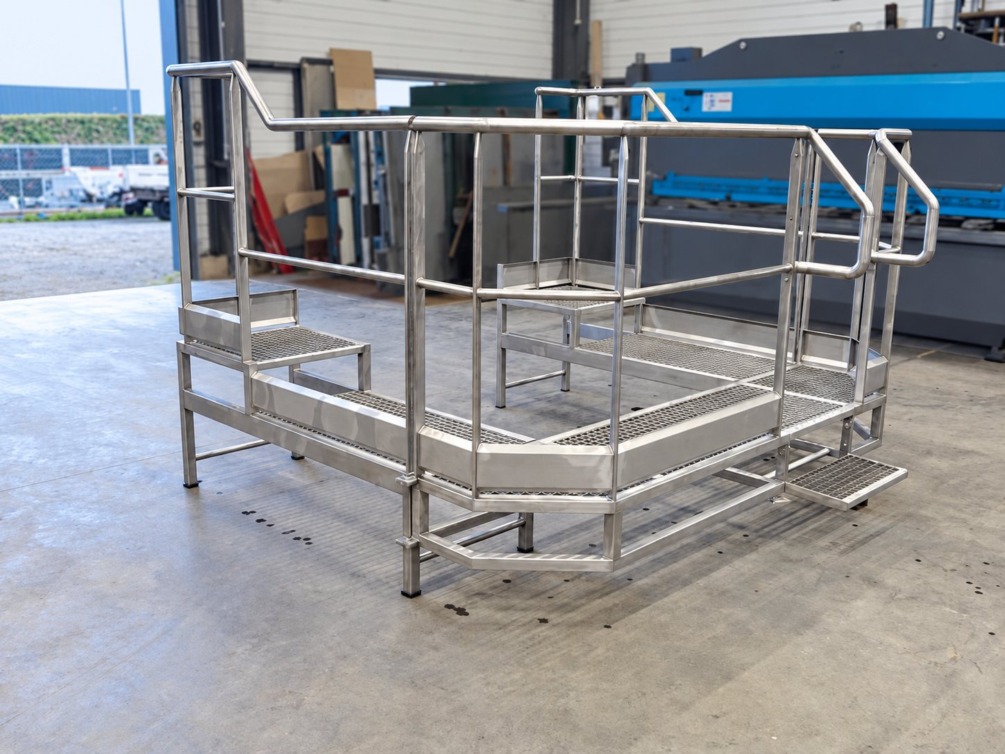 Une plateforme en acier galvanisé avec garde-corps, caniveaux grillagés et petites marches est posée sur le sol en béton d'un atelier de métallerie.