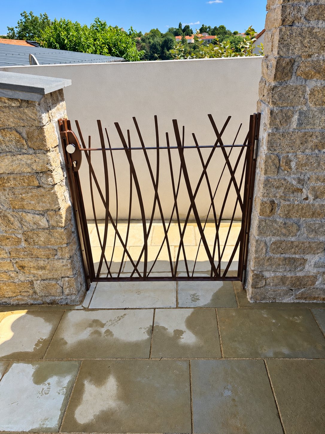 Un portillon en métal brun aux barreaux courbes évoquant des herbes, installé entre deux piliers en pierre donnant sur une terrasse dallée.