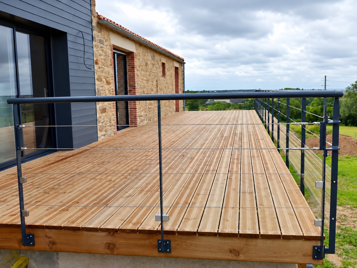 Terrasse en bois entourée d’un garde-corps métallique à câbles inox, devant une maison en pierre.