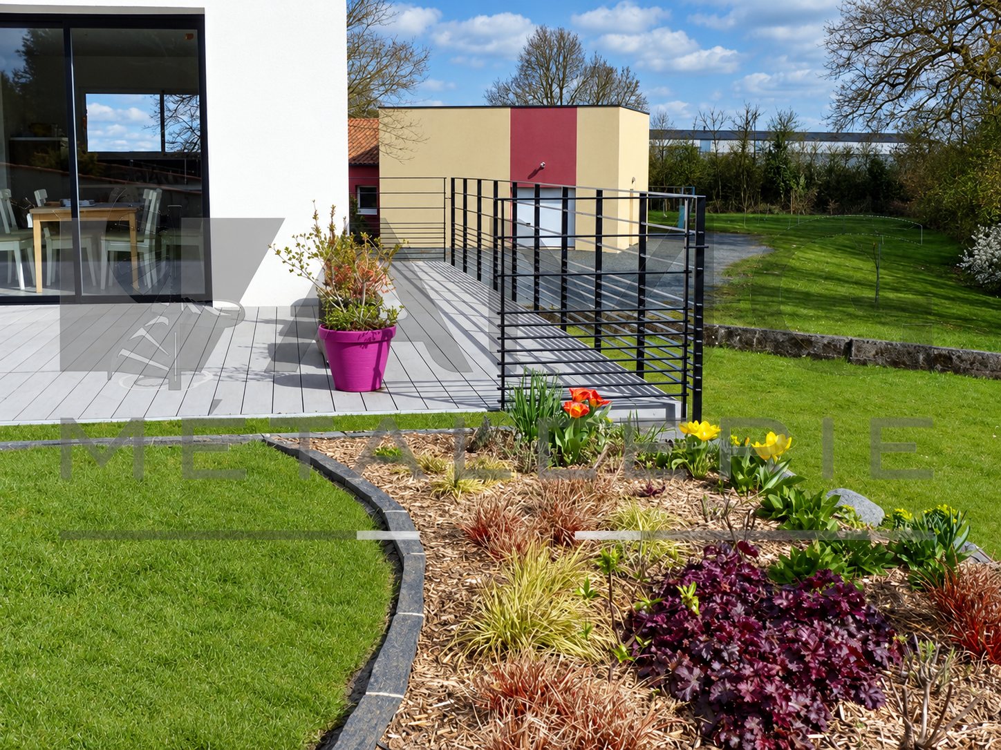 Terrasse en lames grises bordée d'un garde-corps métallique à barreaux horizontaux noirs, avec un pot rose et un massif de fleurs au premier plan.