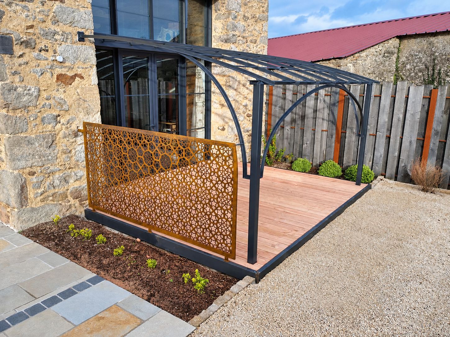 Une terrasse en bois couverte d'une pergola en métal noir, bordée d'un panneau décoratif ajouré en acier rouillé, devant une maison en pierre.