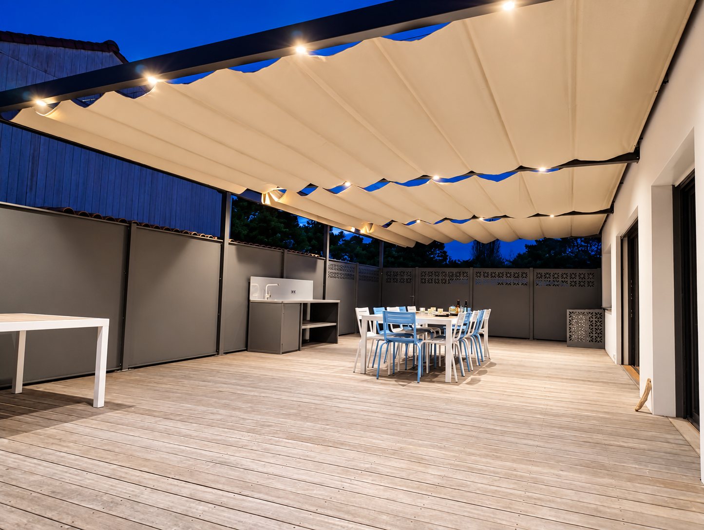 Une grande terrasse en bois au crépuscule, couverte d'une pergola à toile beige rétractable avec spots lumineux, une table et des chaises au fond.