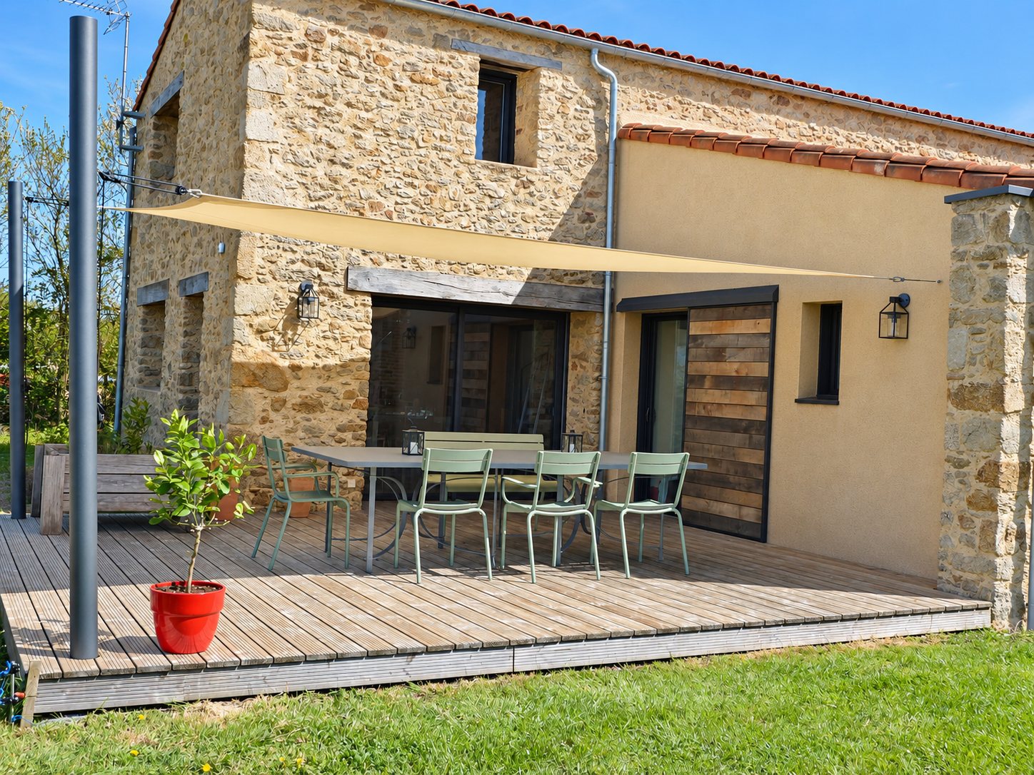 Une terrasse en bois équipée d'une voile d'ombrage beige tendue sur des mâts, avec une table et quatre chaises vertes devant une maison en pierre.