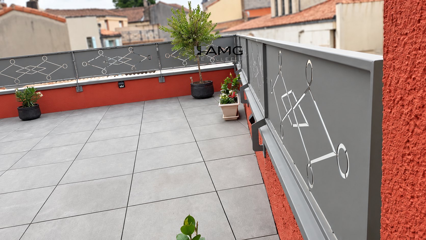Terrasse en dalles grises entourée d'un garde-corps métallique gris à motifs géométriques découpés, avec des plantes en pots et des murs orange.