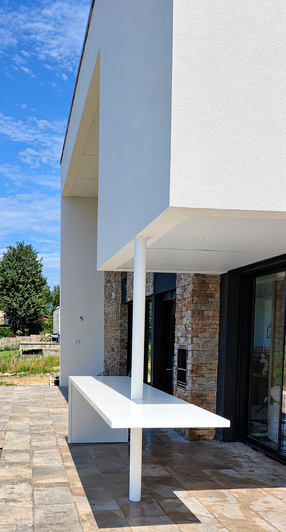 Terrasse dallée d'une maison contemporaine blanche avec un grand plan de table blanc soutenu par un poteau et un mur en pierre apparente.