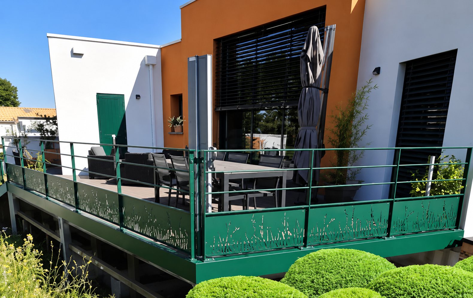 Une terrasse surélevée avec un garde-corps métallique vert, une table et des chaises, devant une maison aux façades orange et blanche, avec des buis arrondis au premier plan.