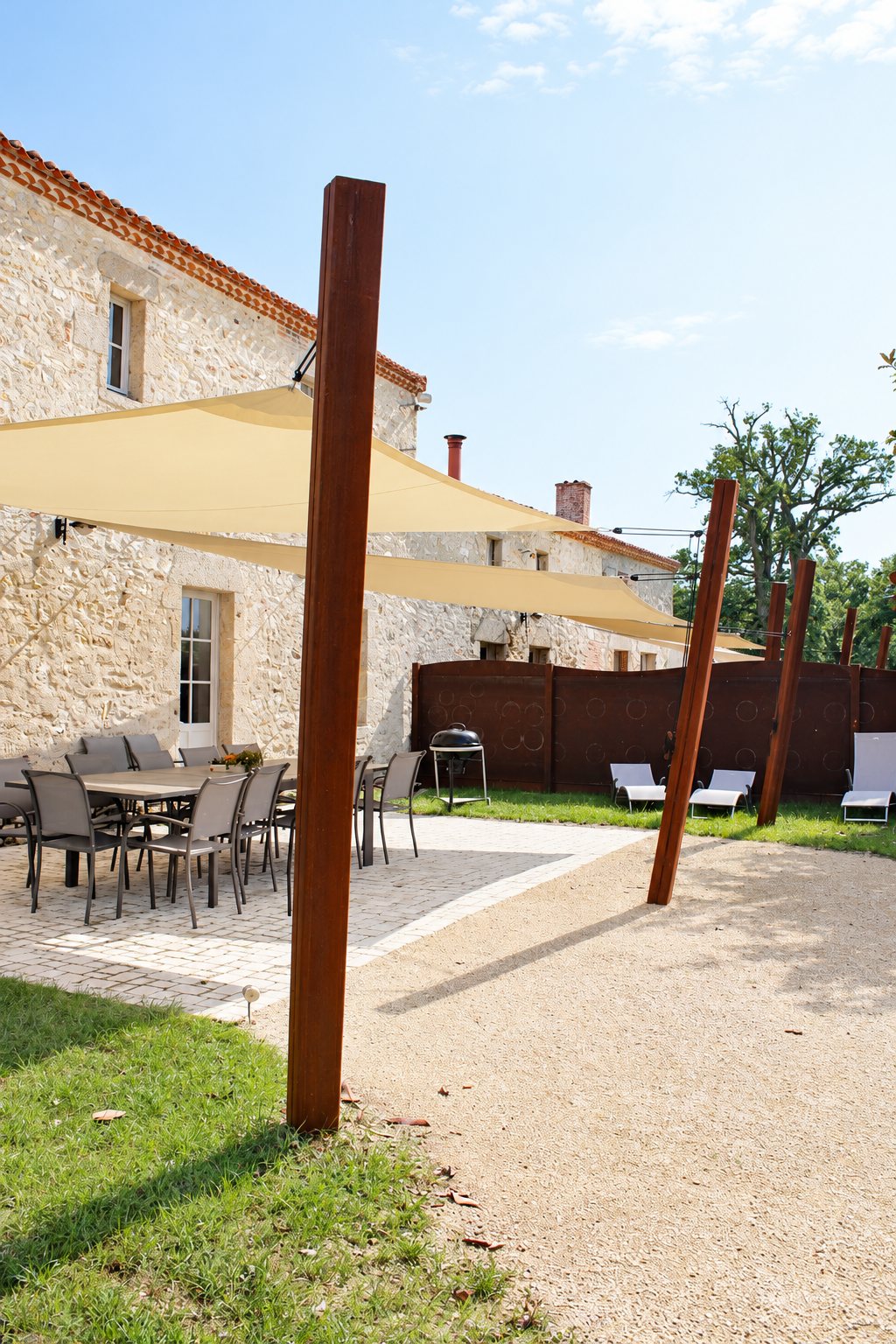 Une terrasse d'une maison en pierre, équipée de grandes voiles d'ombrage beiges fixées à des mâts en acier rouillé, avec une table, des chaises et des transats.